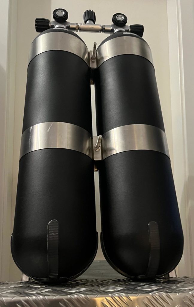 Zwei schwarze Tauchflaschen mit Metallbändern und Druckreglern, auf einem Tisch platziert.